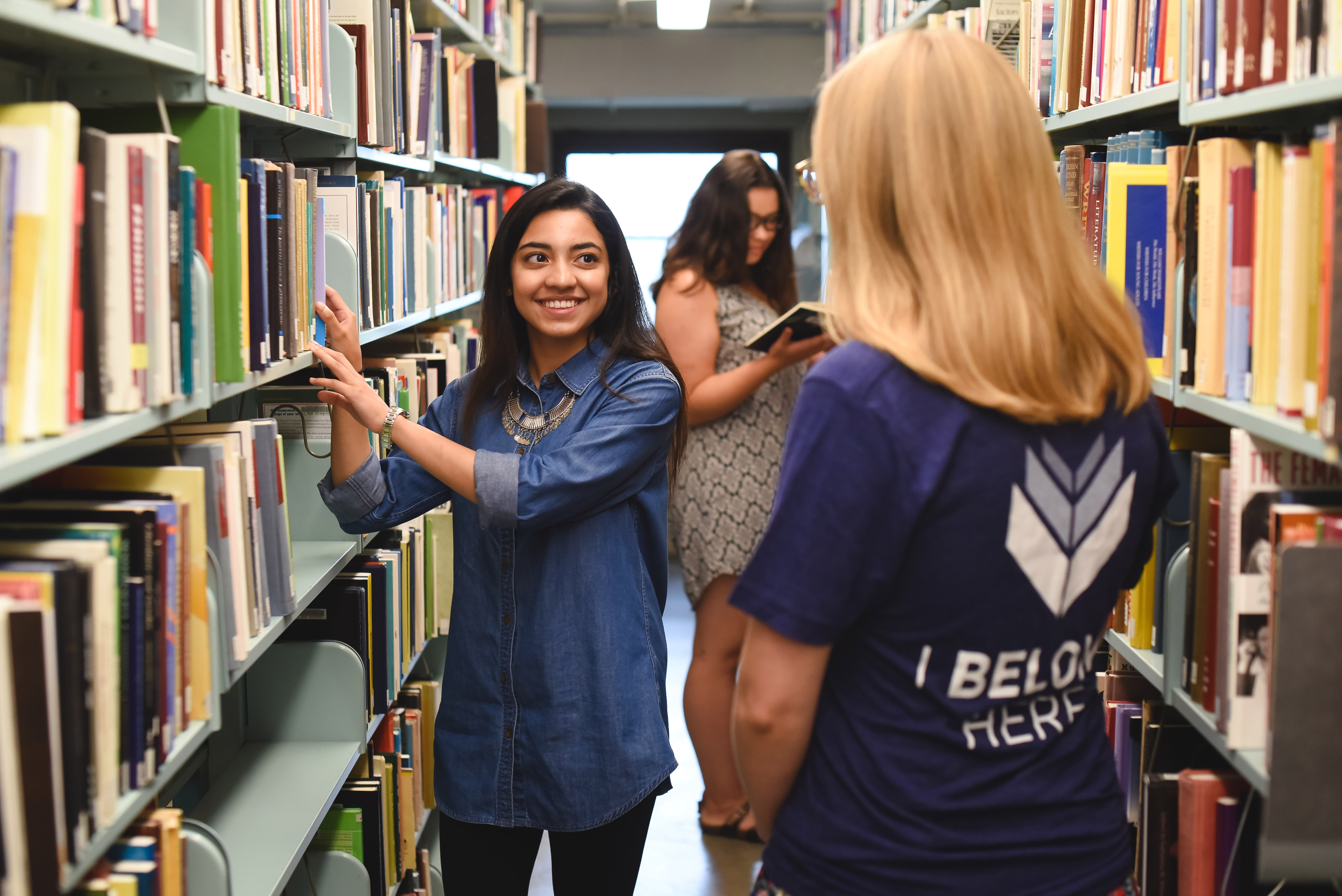 Student pulling book off bookshelf while talking to other subject and another student browsing in the background